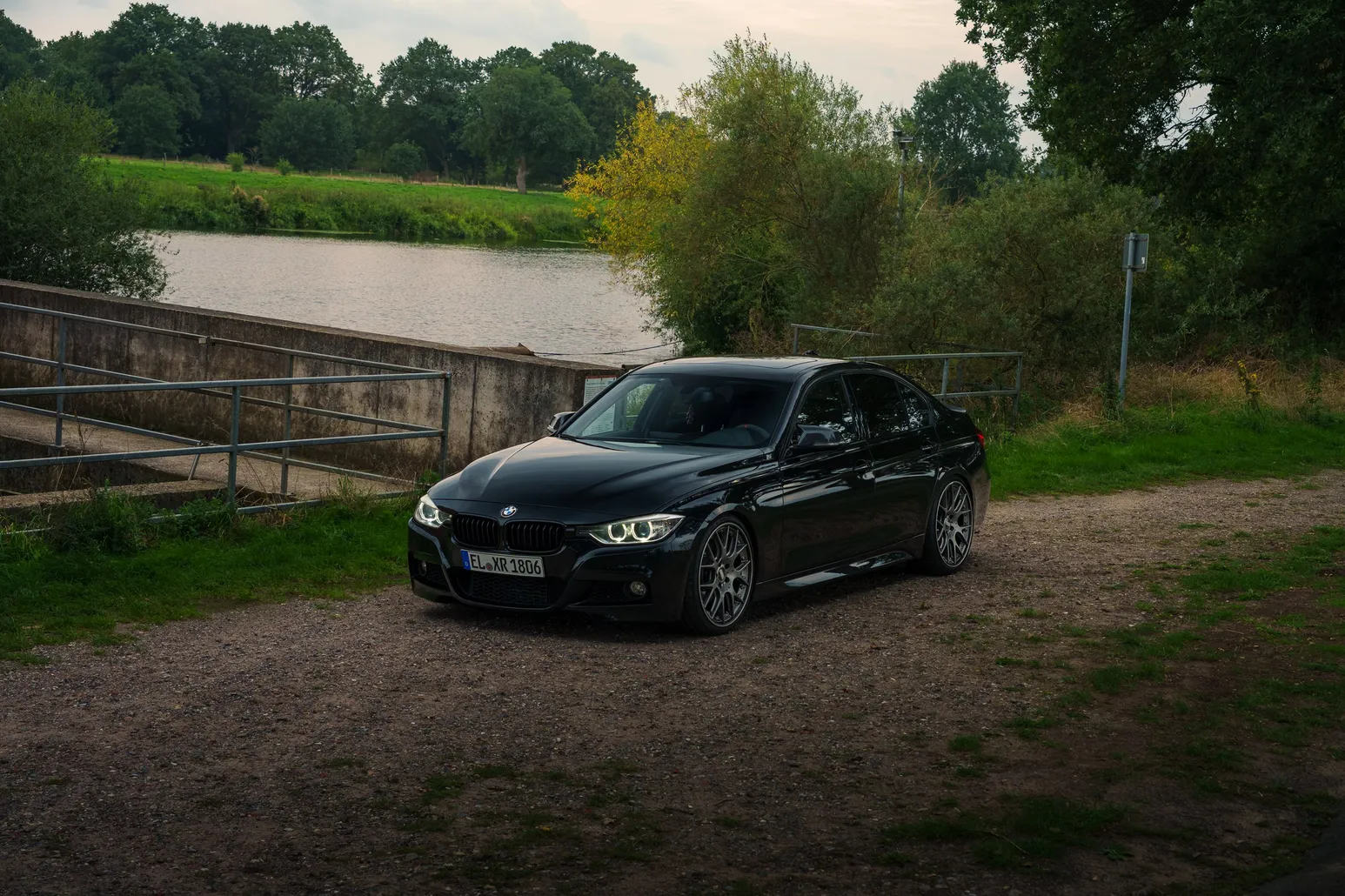 Black BMW F30 by the river with headlights on