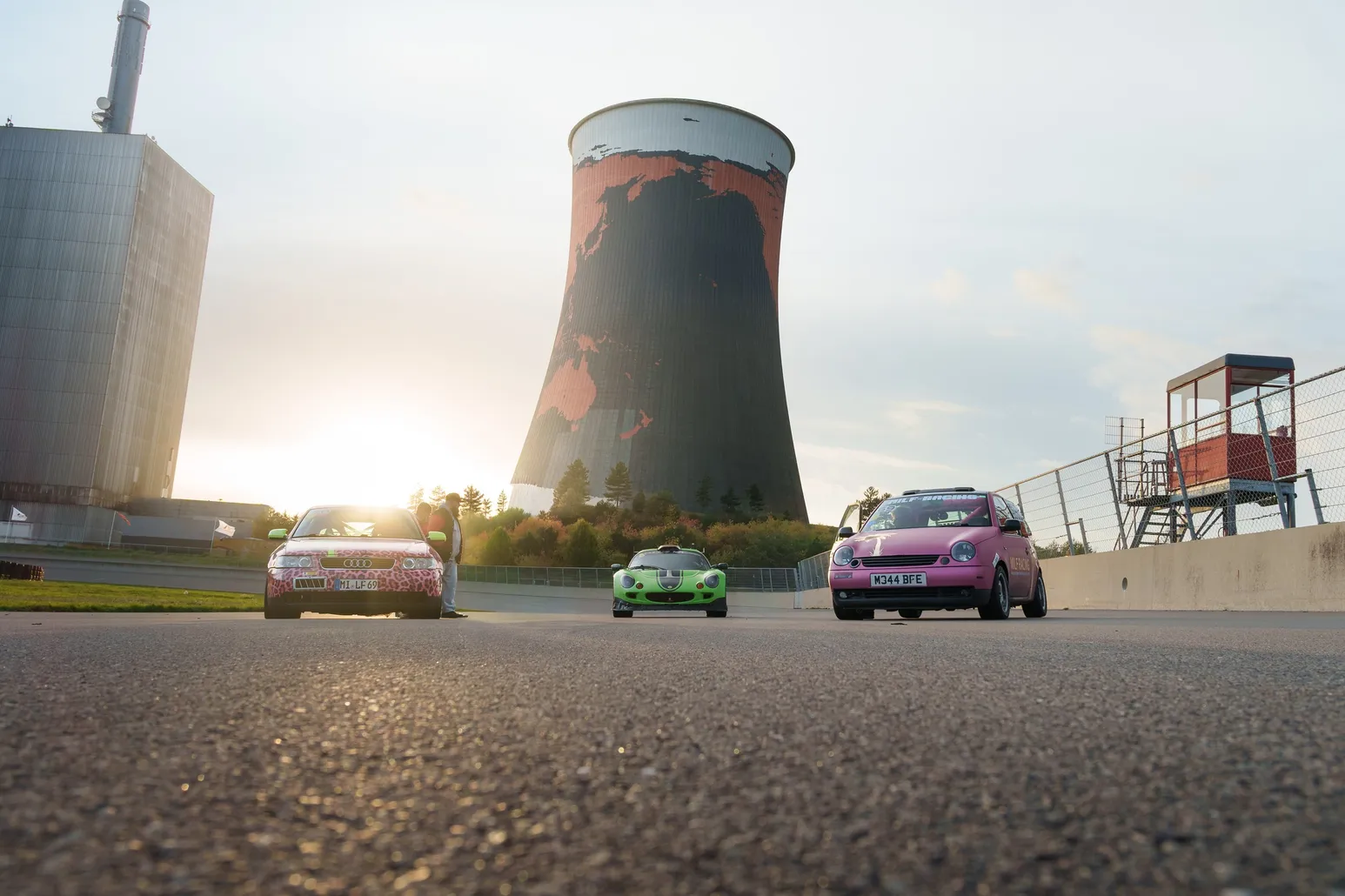 Ground-level view across the asphalt towards three cars in front of the cooling tower
