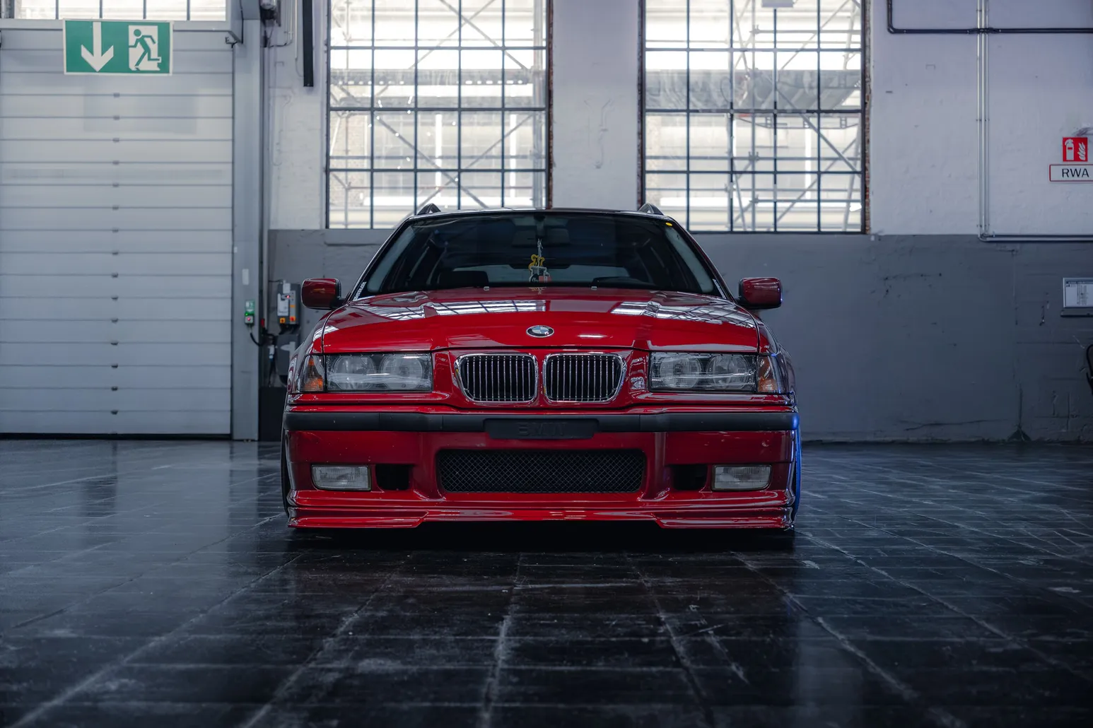 Red BMW E36 head-on in front of large factory windows