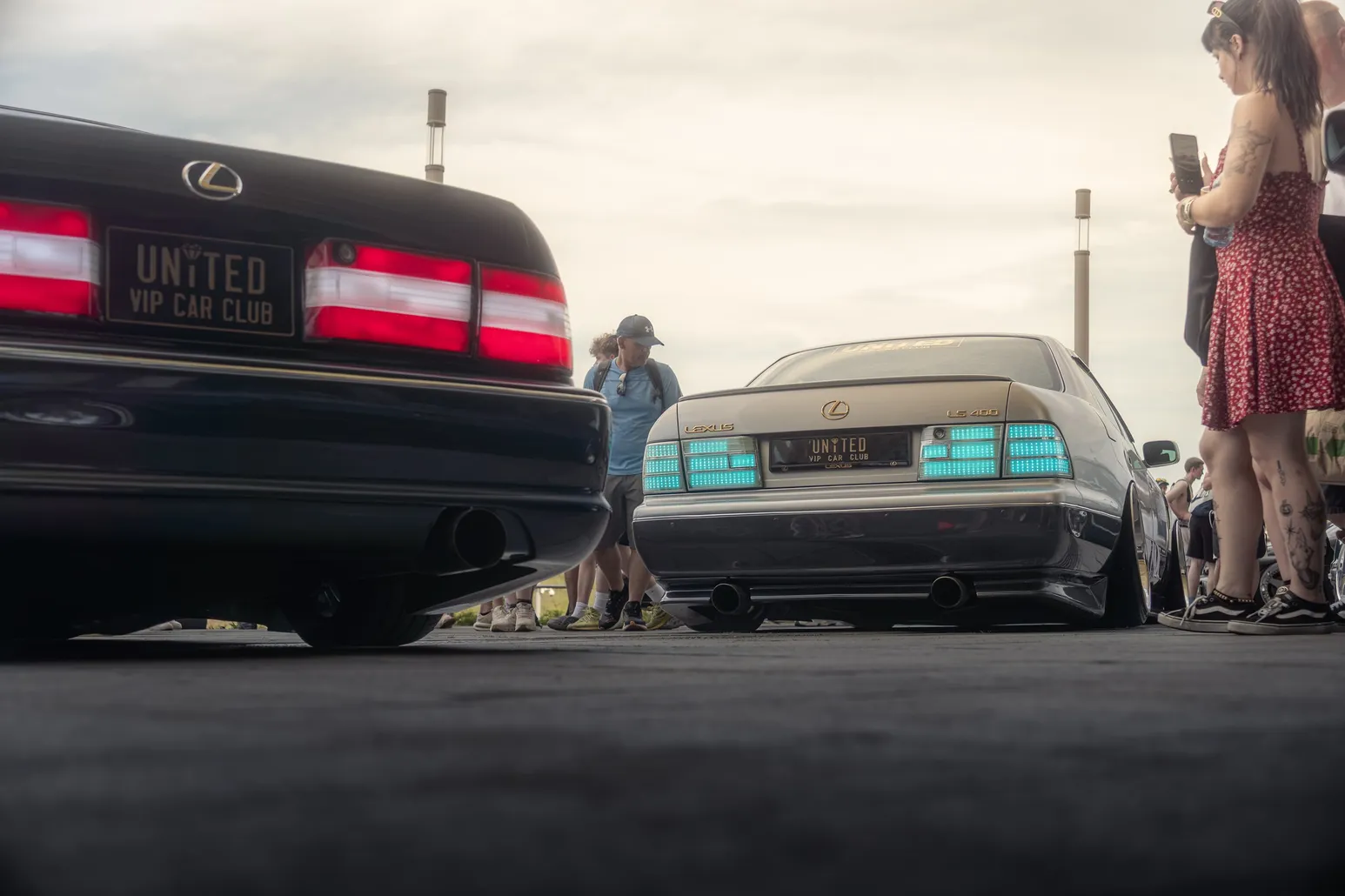Rear of a black Lexus with a club plate, a classic car behind