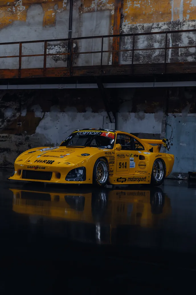 Yellow Porsche race car in front of a rusty hall wall, portrait format